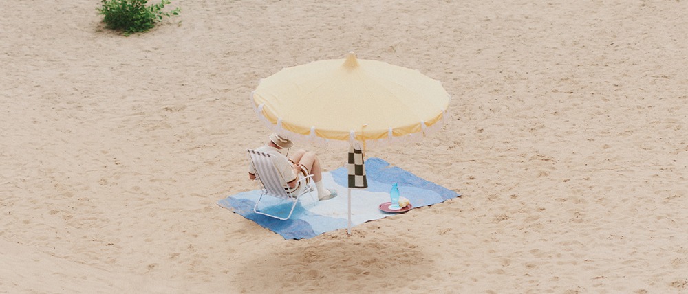 A person sitting on a striped beach chair under a large cream-colored umbrella on a sandy beach, with a blue-hued Urvi rug beneath the chair. Nearby are a water bottle and a striped container.