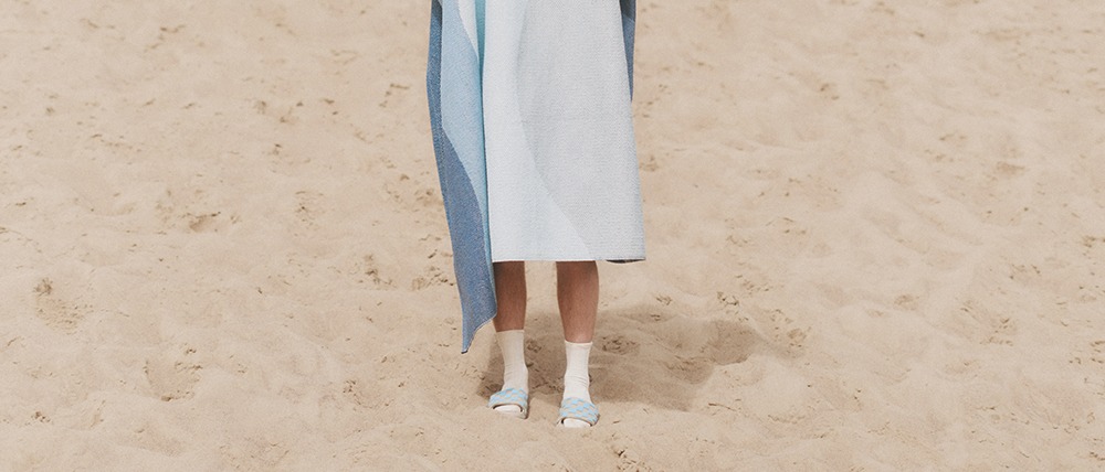 A person standing on the sand wrapped in a blue-hued rug named Urvi, wearing white socks and blue sandals.