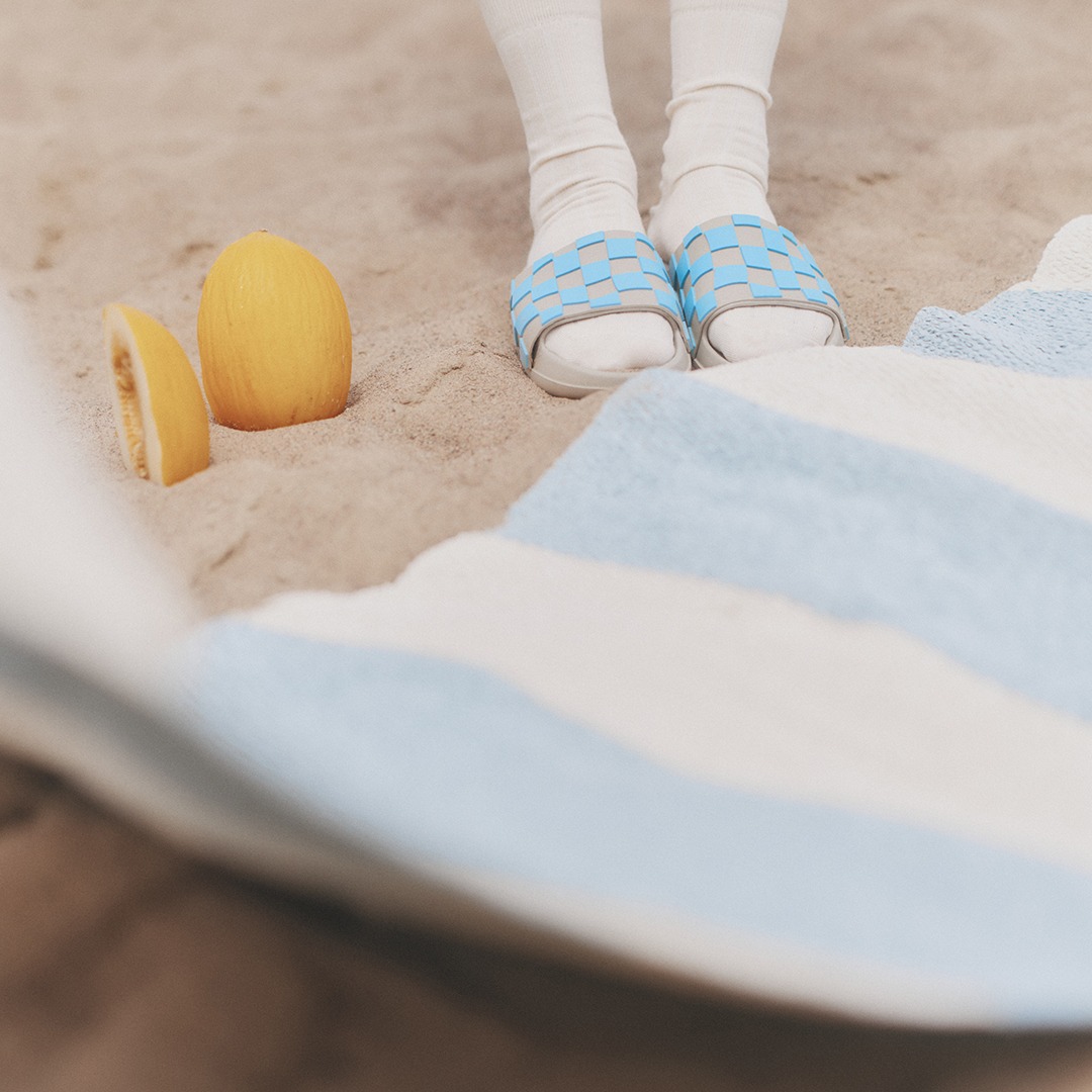 Close-up of feet in blue and white sandals standing on sand, next to a blue and white striped rug named Bob. An orange and a slice of orange are also placed in the sand.