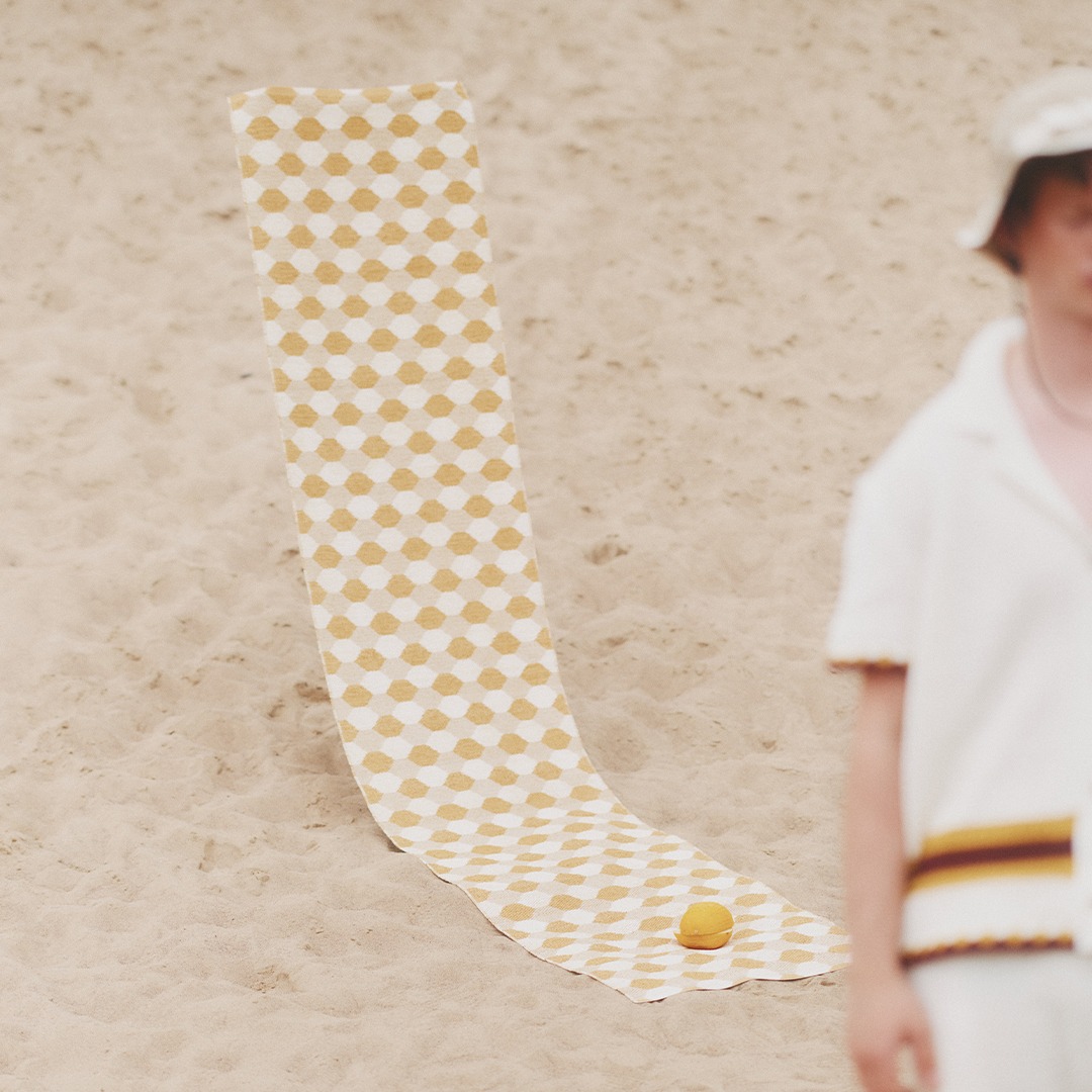 A long, yellow and white checkered rug named Dana laid out on the sand, with a whole orange placed near the bottom. A person wearing a white outfit is partially visible on the right side.