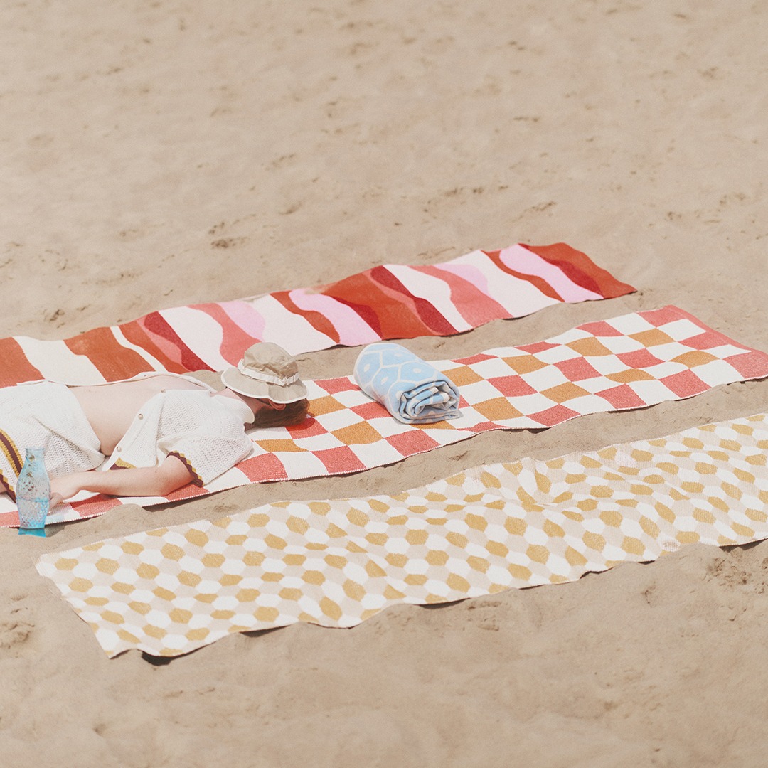 The image shows three rugs on the sand. The furthest rug is an Urvi in red hues, the middle rug is a Pix with a flamingo pink square pattern, and the closest rug is a Dana in ochre. On the middle rug, there is also a Honey pattern blanket next to a person lying down.